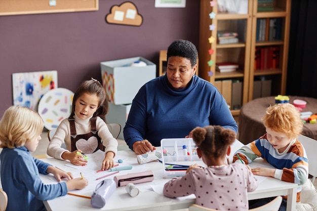 Mature african american teacher sitting among group of intercultural diligent little learners intera