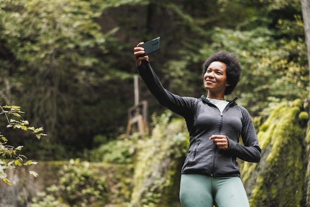 Donna afroamericana matura che fa una videochiamata mentre si trova vicino alla cascata e si gode il panorama durante la sua escursione in montagna.