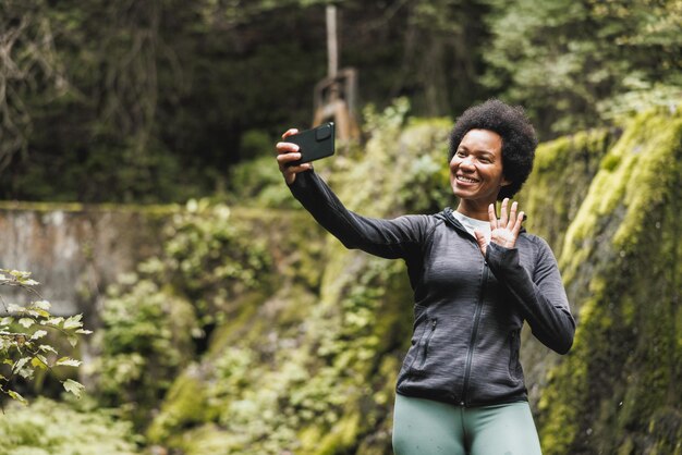 성숙한 아프리카계 미국인 여성이 폭포 근처에 서서 산에서 하이킹하는 동안 경치를 즐기면서 셀카를 찍습니다.