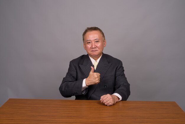 Photo mature asian businessman sitting with wooden table against gray