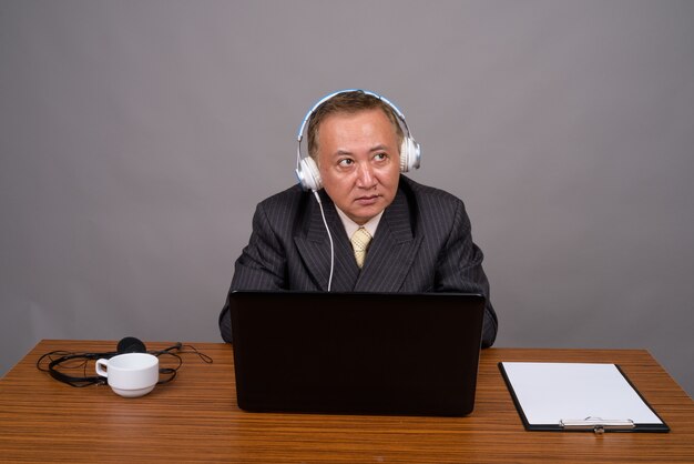 Mature Asian businessman sitting with wooden table against gray