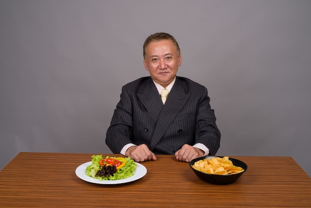 Mature Asian businessman sitting with wooden table against gray