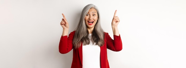 Mature asian businesswoman with grey hair wearing red blazer and pointing fingers up smiling surpris