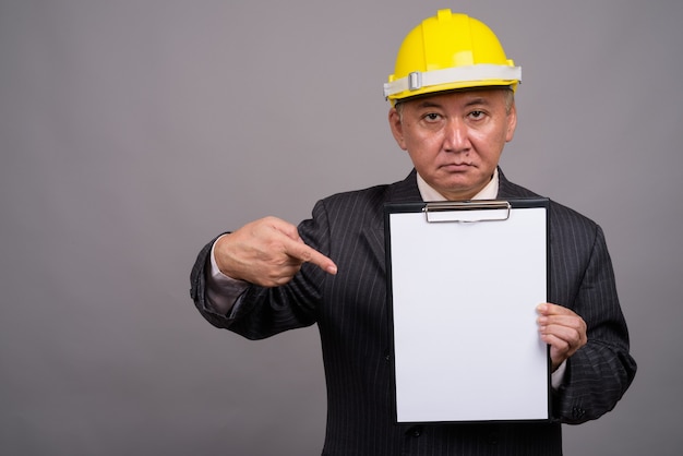 Зрелый азиатский строительный бизнесмен против серой стены