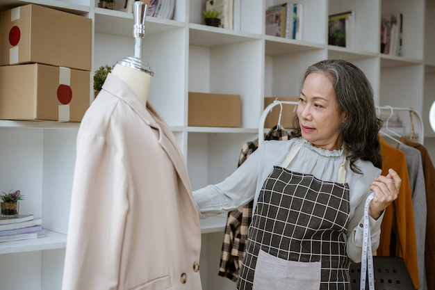A mature Asian female tailor measuring a suit on a mannequin with a measuring tape