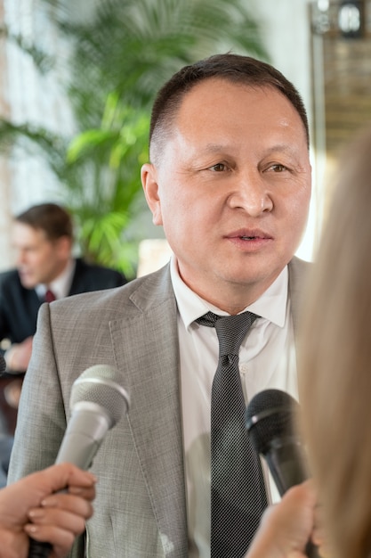 Mature Asian male delegate in formalwear standing in front of journalists with microphones and answering their questions during interview