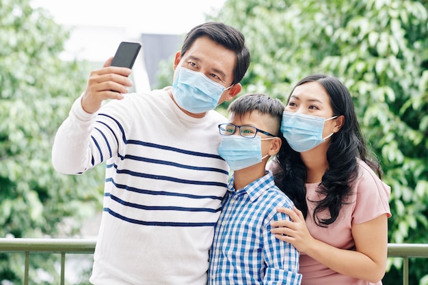 Mature Asian man wearing medical mask due to coronavirus pandemic and taking selfie with wife and preteen child