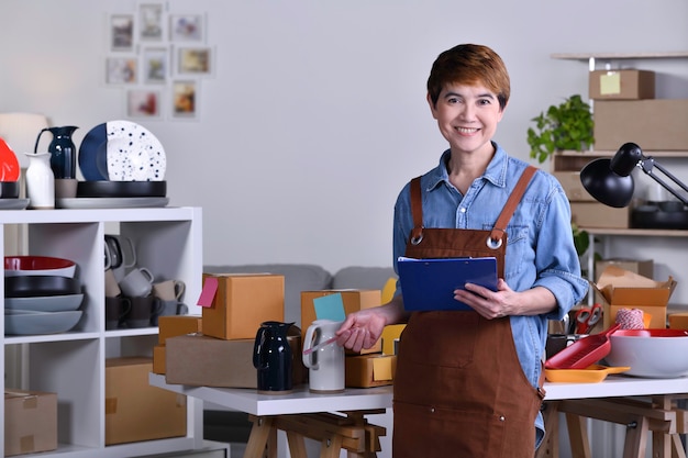 Foto imprenditrice asiatica matura, titolare di un'azienda in piedi di fronte al suo prodotto in ceramica di argilla e lavora a casa