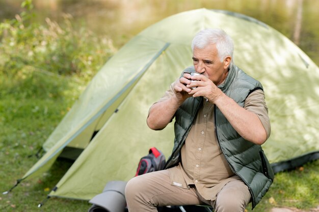 Mature backpacker with tourist mug sitting by tent and having hot drink and rest in the forest