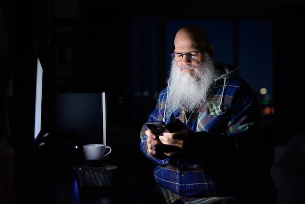 Mature bald bearded hipster man using phone at home late at night in the dark