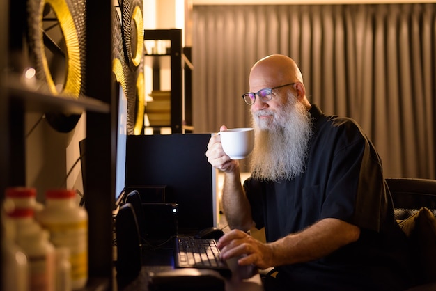 Mature bald bearded man drinking coffee while working overtime at home late at night