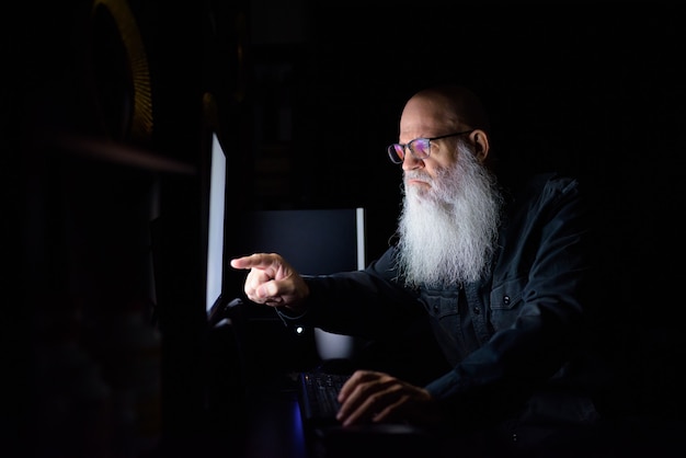 Mature bald bearded man working overtime at home in the dark