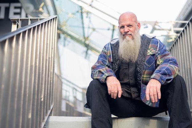 市内の歩道橋に座っている長いひげを持つ成熟したハゲヒップスター男