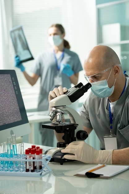 Mature bald male in protective eyewear, mask and gloves studying new vaccine in microscope against female assistant with x-ray image