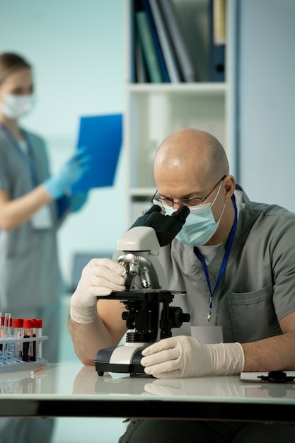 Mature bald male scientist in protective mask and gloves studying chemical samples in microscope against female assistant working with documents