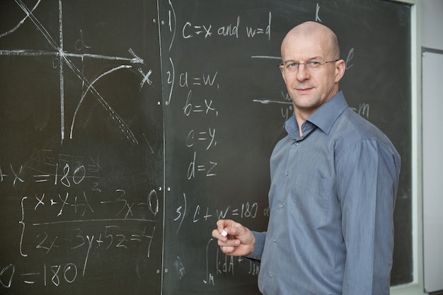 Mature bald professor in smart casualwear holding piece of chalk while standing by blackboard in classroom or lecture hall and looking at you