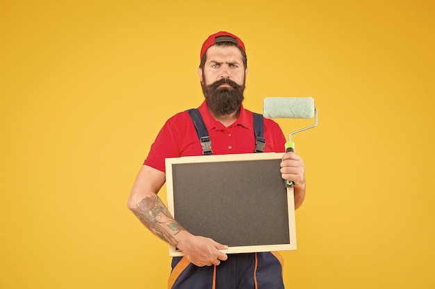 Mature bearded guy painter worker in boiler suit uniform using paint roller tool for painting wall and hold blackboard for copy space advertisement advertising