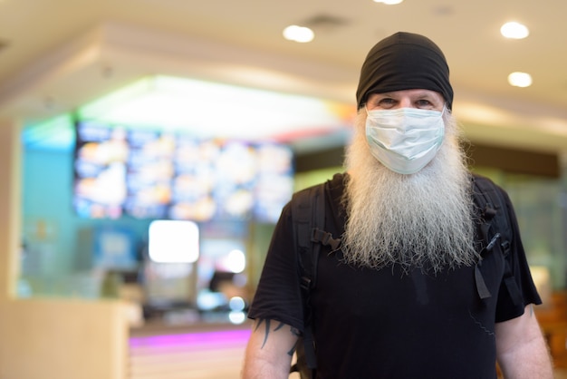 Mature bearded hipster man as backpacker wearing mask inside the mall