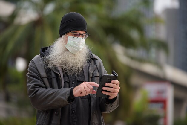 屋外の電話を使用してマスクを持つ成熟したひげを生やした流行に敏感な男