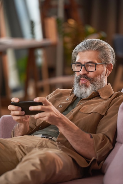 Mature bearded man in eyeglasses resting on sofa and playing on his smart phone at home