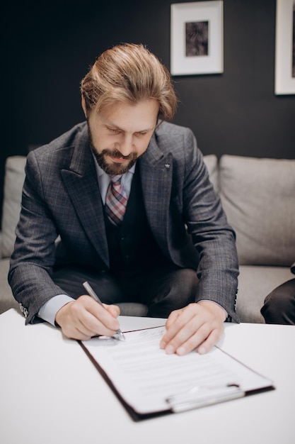 Mature bearded man signing documents while sitting on couch