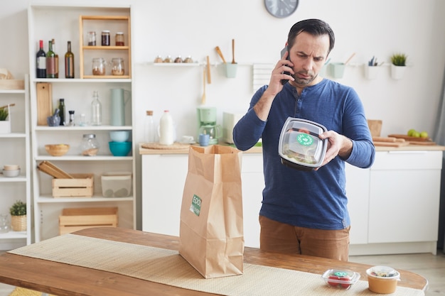 mature bearded man speaking by smartphone while unpacking food delivery bag in kitchen interior copy space