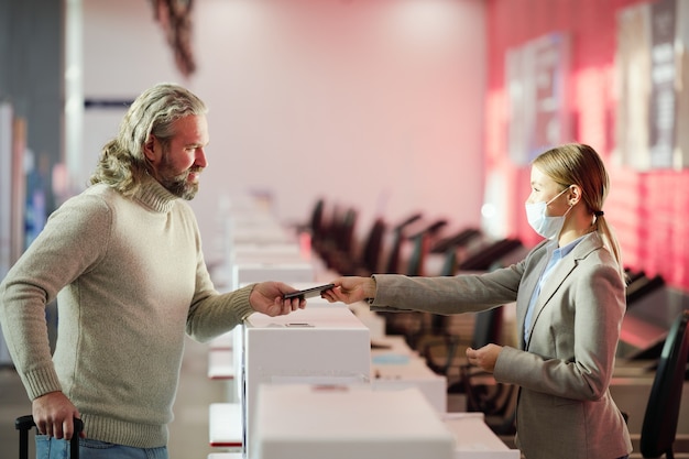 Mature bearded man standing at reception counter and taking back his passport after registration before flight at the check desk