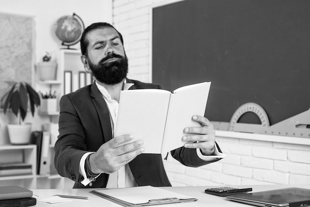 Зрелый бородатый учитель в костюме сидит в школьном классе с доской и читает книжную литературу