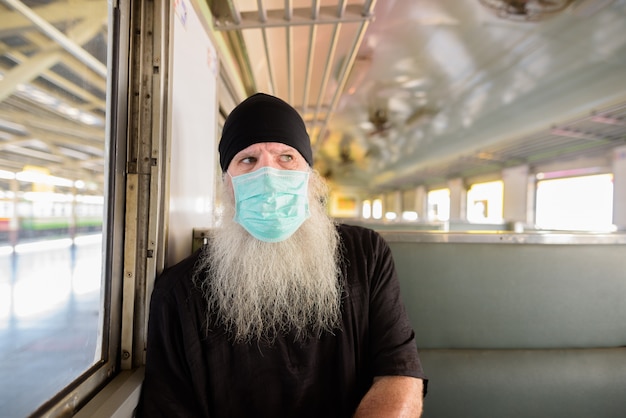 Mature bearded tourist man thinking with mask for protection from corona virus outbreak riding the train