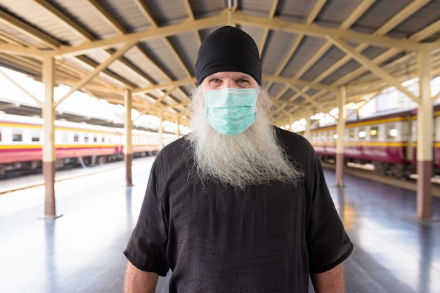 Mature bearded tourist man with mask for protection from corona virus outbreak at the railway station