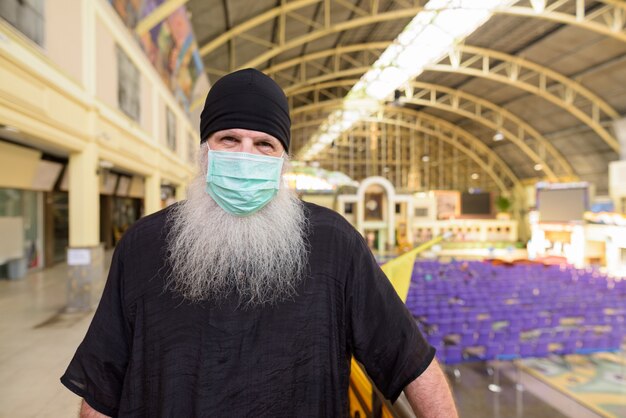 Mature bearded tourist man with mask for protection from corona virus outbreak social distancing at railway station