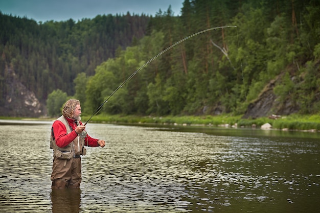 mature bearded whire wet man is standing in water in middle of river and fishing flyfishing, eco tourism.