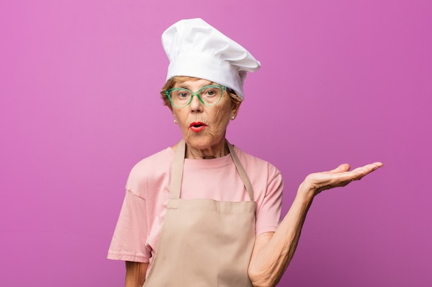 Mature beautiful old woman looking surprised and shocked, with jaw dropped holding an object with an open hand on the side