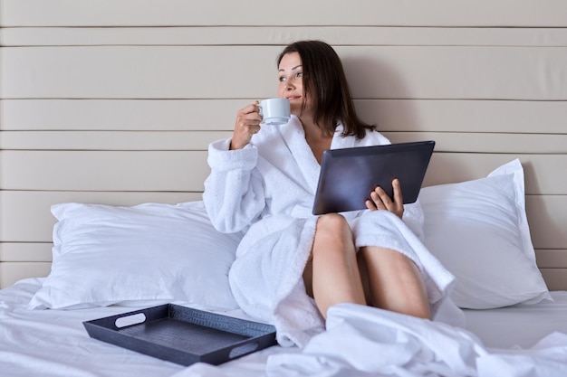 ホテルで休んでいる成熟した美しい女性。ベッドに座って、デジタルタブレットを見て楽しんでコーヒーのカップと白いバスローブを着た女性