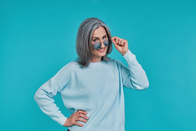 Mature beautiful woman looking at camera and smiling while standing against blue background