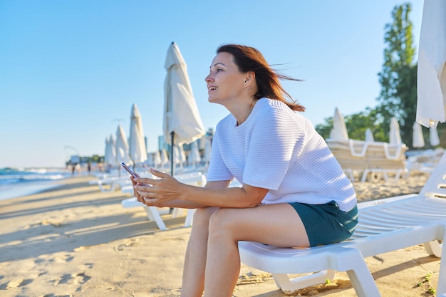 夕暮れ時のスマートフォンを使用して海のビーチに座っている成熟した美しい女性