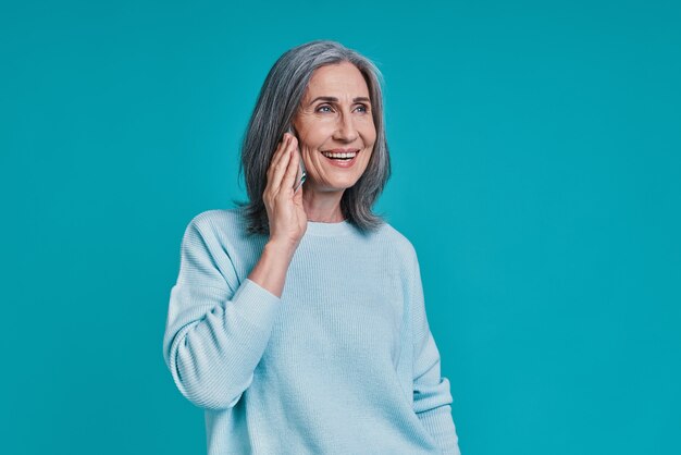 Mature beautiful woman talking on smart phone and smiling while standing against blue background