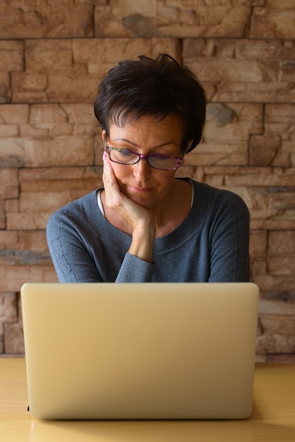 Mature beautiful woman using laptop while wearing eyeglasses and resting chin on hand