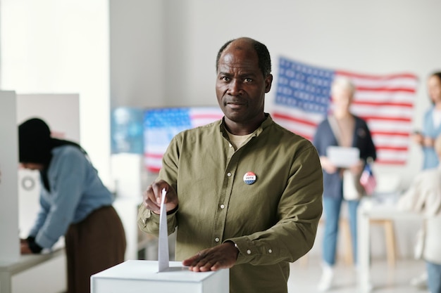 Фото Зрелый темнокожий мужчина смотрит в камеру, опуская бумагу в урну для голосования