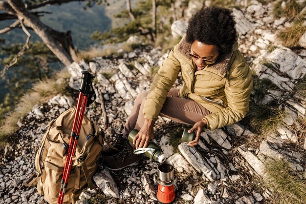 Mature black woman sitting on the top a hill and having tea break during her hike in the mountains.