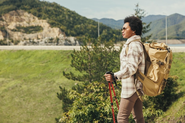 Зрелая чернокожая женщина с рюкзаком и треккинговыми палками в одиночестве путешествует по горе в прекрасный солнечный осенний день.