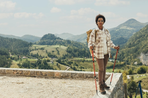 Mature black woman with backpack and trekking poles standing in nature and enjoying in fresh air while hiking on mountain.