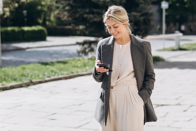 Mature blond business woman in a gray jacket smiling walking around the city and holding a phone