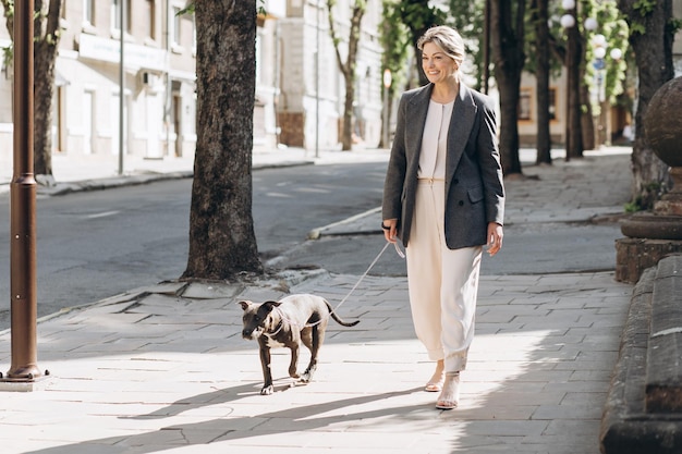 Зрелая блондинка-деловая женщина в серой куртке улыбается, выгуливая свою собаку породы амстафф и разговаривая по телефону