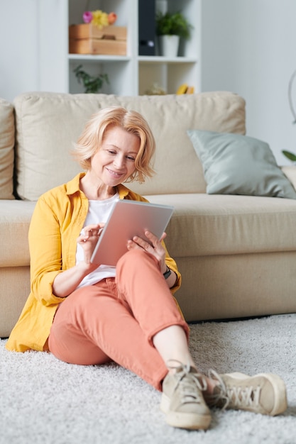 Mature blond relaxed businesswoman with mobile gadget sitting on the floor by couch in living-room while planning work during quarantine