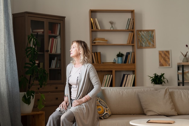 Mature blond restful woman in grey casualwear sitting on couch in living-room in front of camera against shelves and cabinet with books