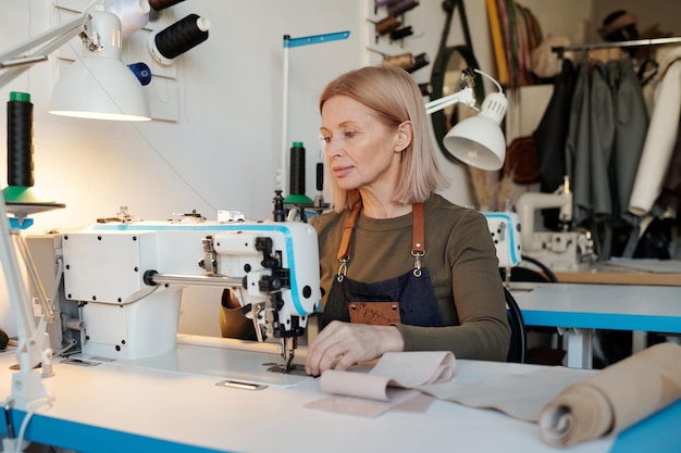 Mature blond seamstress sitting by electric sewing machine during work