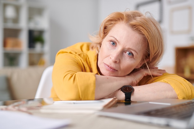 Donna di affari isolata auto bionda matura che ti guarda mentre giaceva sulla scrivania davanti al computer portatile durante il lavoro a distanza a casa