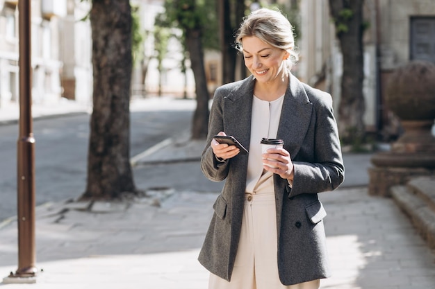 Donna d'affari sorridente bionda matura che cammina per la città bevendo caffè e parlando al telefono in primavera su uno sfondo di fiori lilla