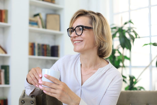 お茶やコーヒーを持っているソファで成熟したブロンドの女性。